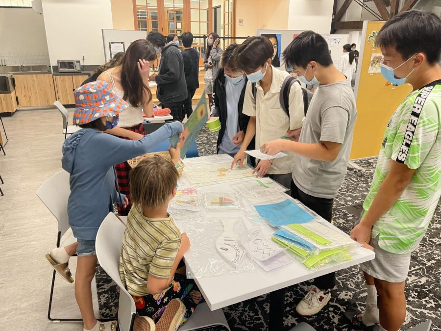 Many students gather around, looking at the artwork created by Dr. Greco’s children. Dr Greco’s children hold up their artwork, showing the Webb students in Hooper. Around the table are Jordan Granda (‘22) and Christopher Chung (‘22) as they look down at the artwork displayed on the table.