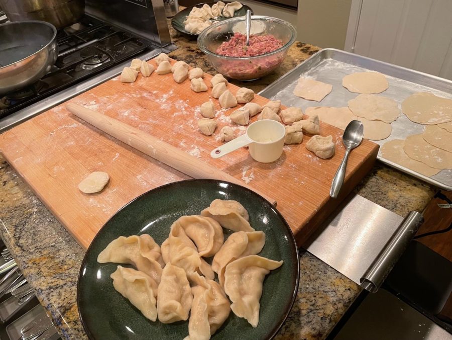 Emily makes dumplings with her family, showing the different steps on her cutting board. The wrappers are being rolled out as small dough balls to form thin circles, the pink filling is ready in the glass bowl, and some boiled dumplings sit on a plate, ready for consumption.