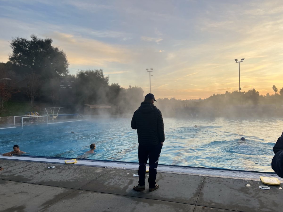 Brian Caldwell, Triathlon team head coach, stands next to the pool as swimmers flip turn and do another lap. The steamy fog reflects a freezing temperature of 35 degrees fahrenheit at 7:00 a.m. in the morning. Despite the cold, swimmers persevere and continue training for their full triathlon by the end of the season, with breathtaking dawns as their constant companion. 